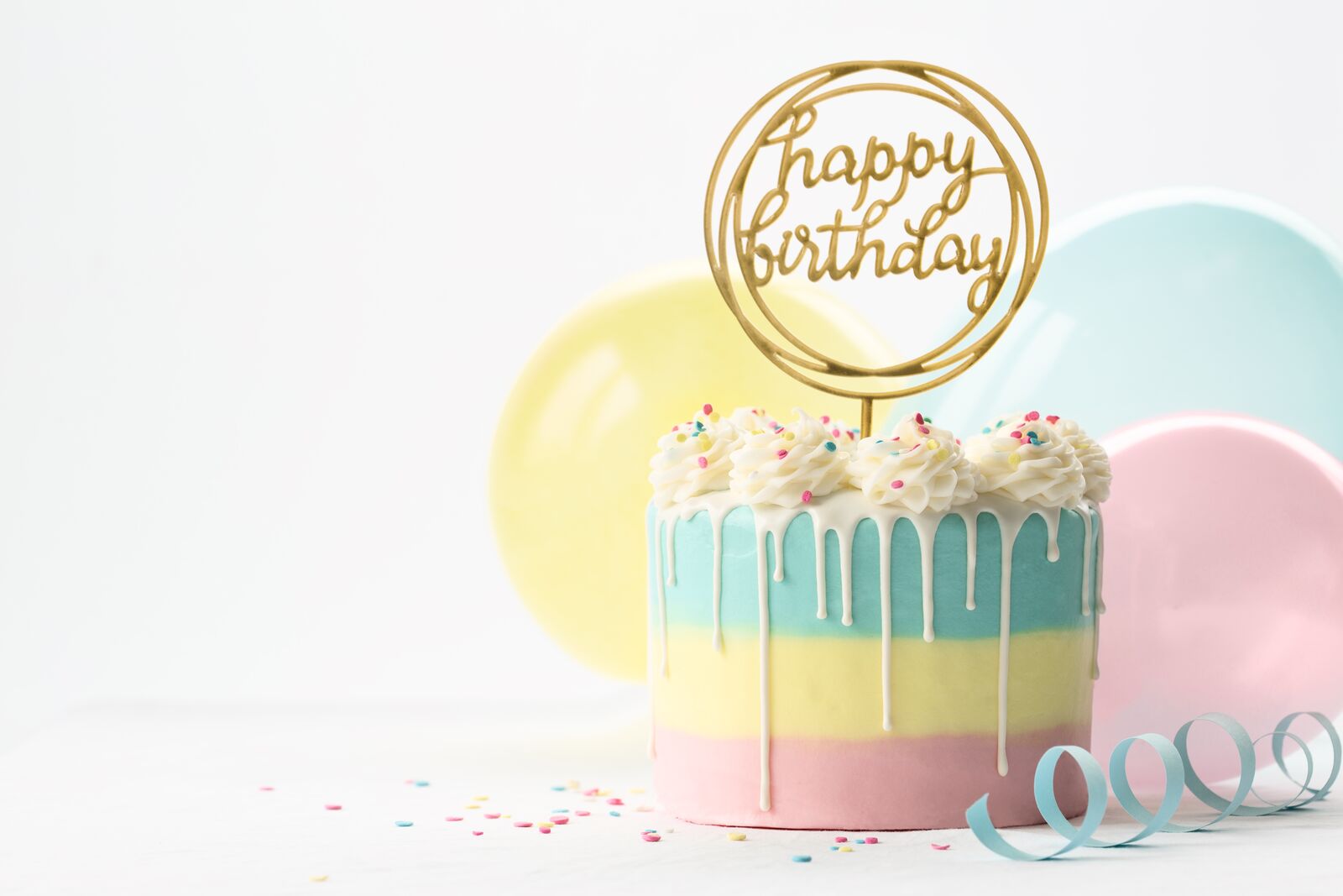 Pastel birthday cake with white cream, sprinkles and balloons.