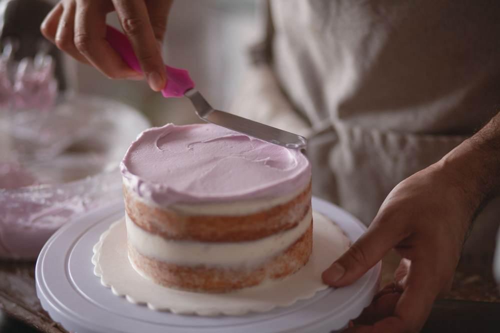 Baker spreading pink frosting on a layered cake using an icing spatula.